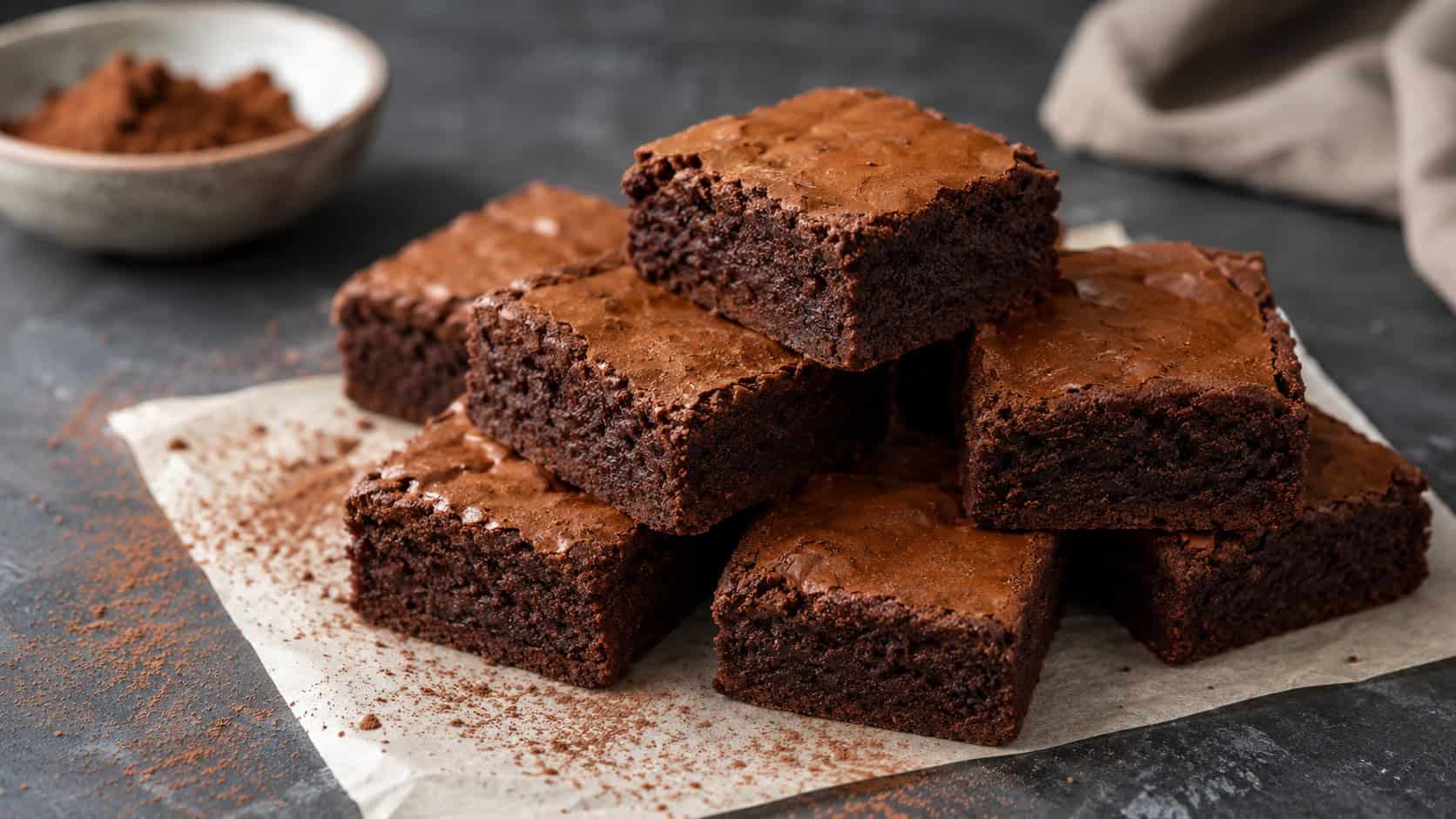 fudgy chocolate brownies cut into neat squares stacked on parchment, shown in a wide horizontal shot on a dark slate surface with a dusting of cocoa, natural light, shallow depth of field, landscape orientation