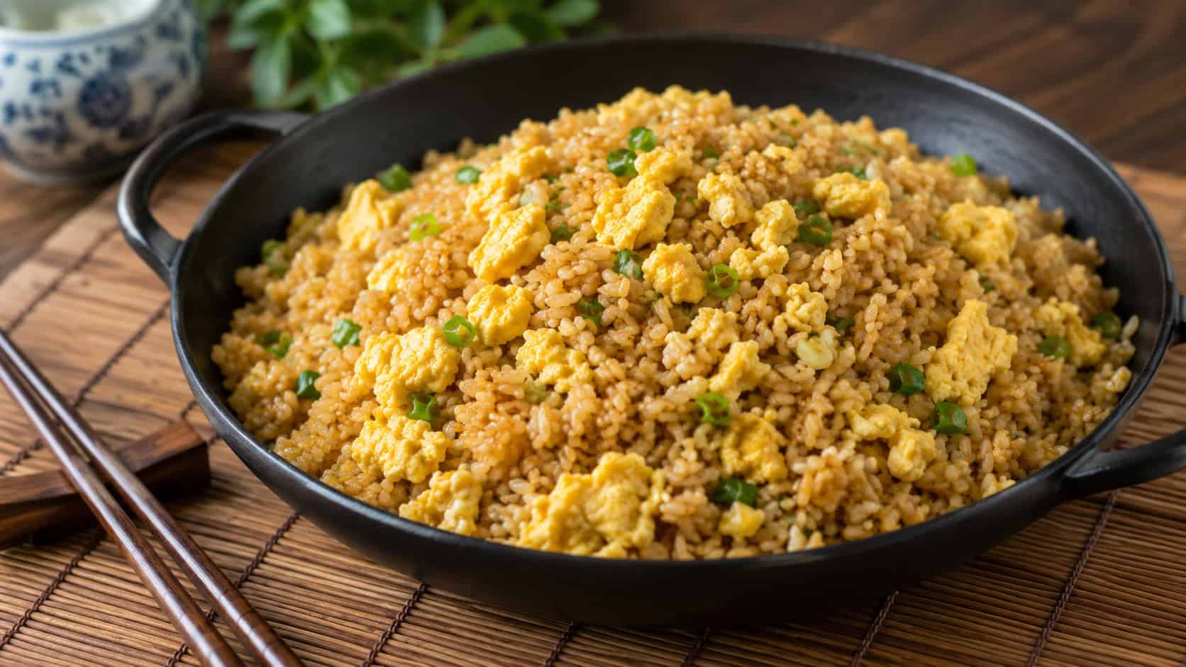 golden egg fried rice flecked with scrambled egg in a dark skillet, shown in a wide horizontal shot on a bamboo mat with chopsticks resting nearby, natural light, shallow depth of field, landscape orientation