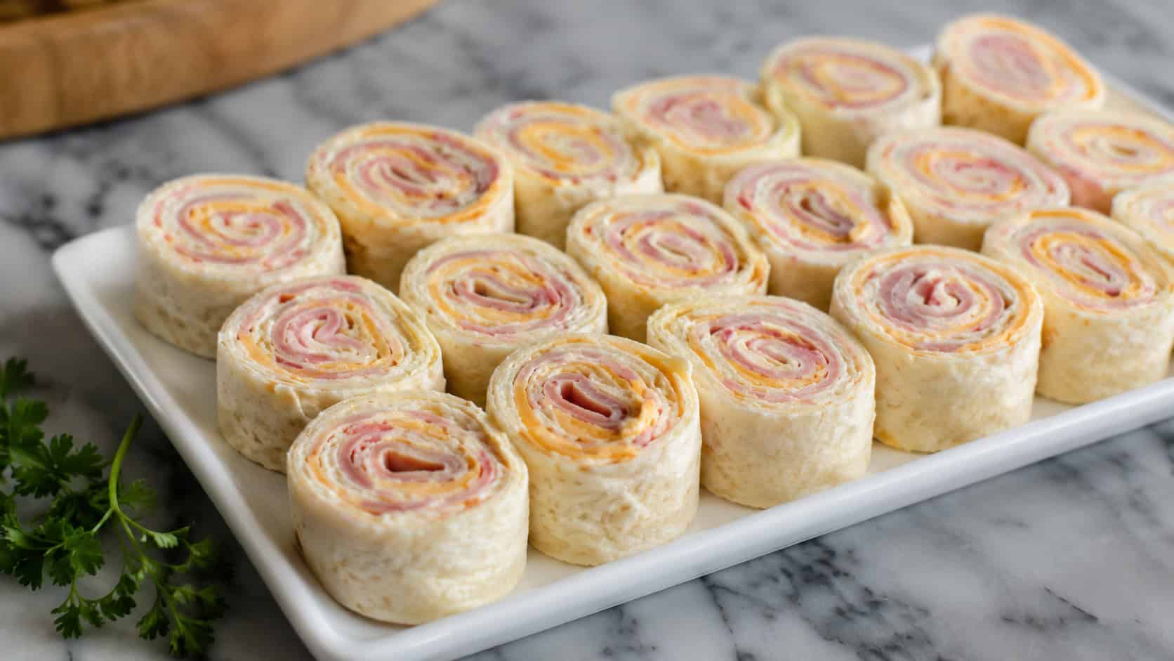 ham and cheese pinwheels arranged cut-side up on a white platter, shown in a wide horizontal shot on a marble counter with a sprig of parsley nearby, natural light, shallow depth of field, landscape orientation