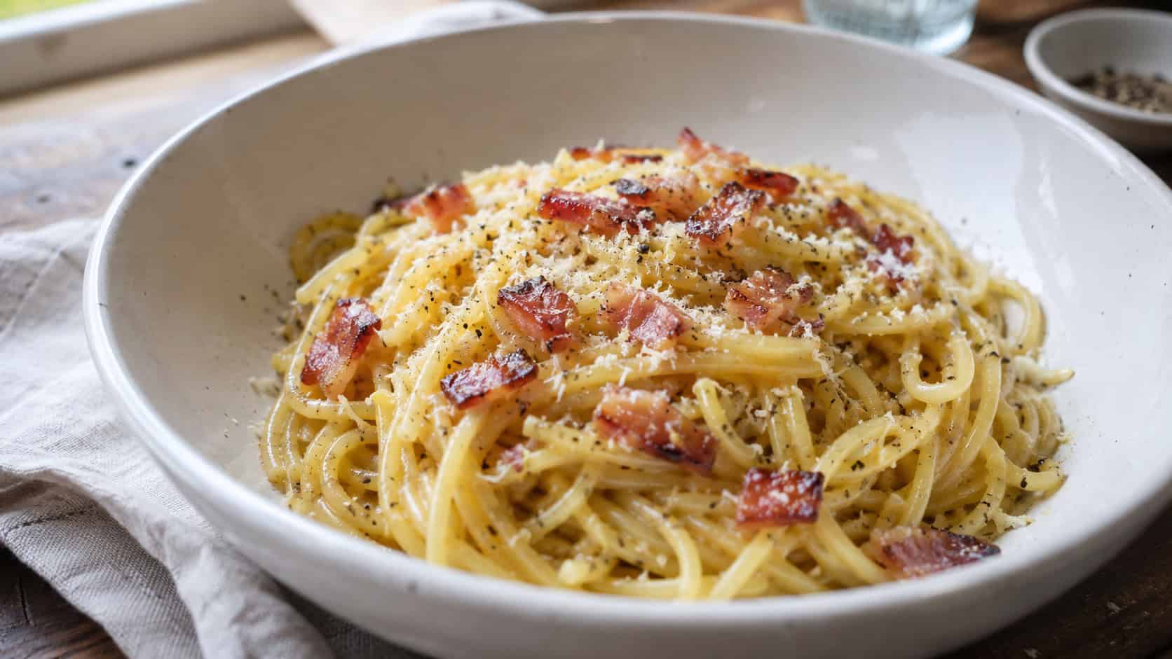 a wide bowl of glossy spaghetti carbonara twirled into a nest, crispy pancetta and cracked black pepper on top, a dusting of grated Parmesan, on a rustic wooden table with a linen napkin, natural window light, shallow depth of field, landscape orientation