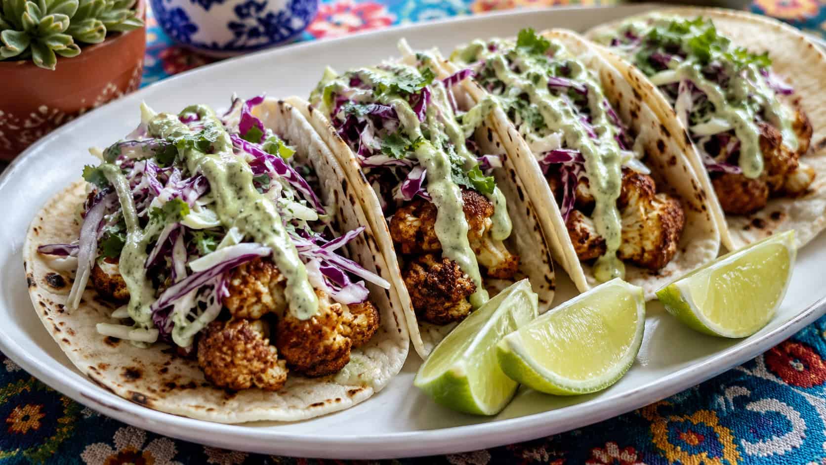 crispy roasted cauliflower tacos topped with shredded cabbage and lime crema on a wide platter with lime wedges, in a wide horizontal shot, on a colorful table, natural light, shallow depth of field, landscape orientation
