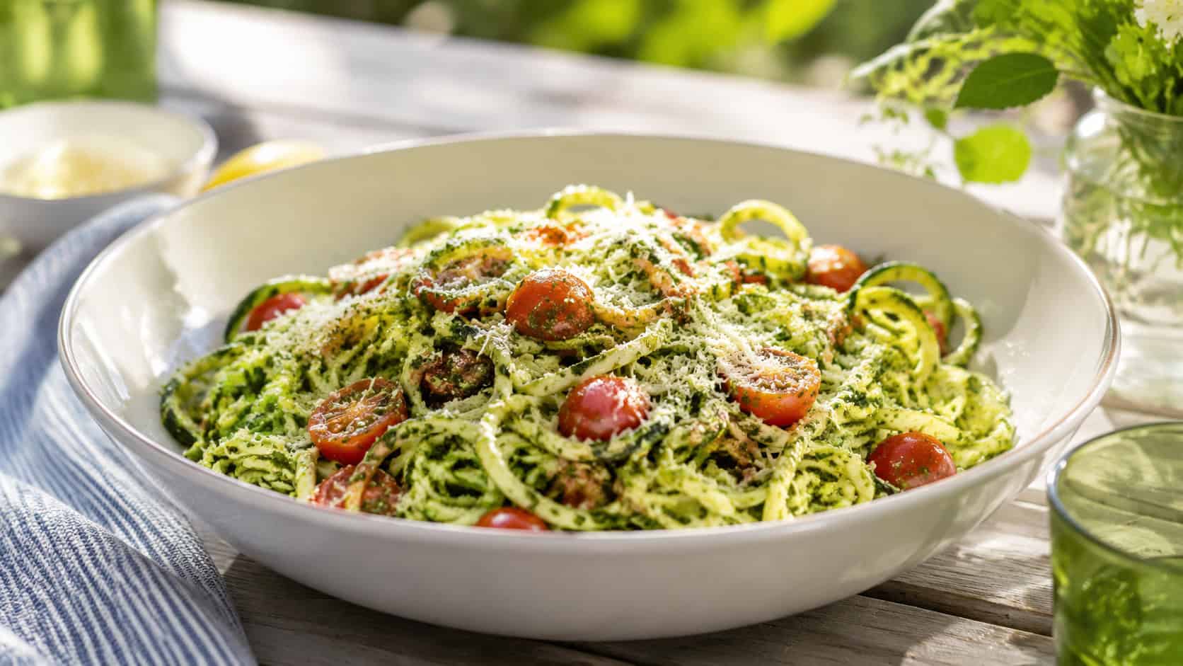 zucchini noodles tossed in green pesto with cherry tomatoes and parmesan in a wide bowl, in a wide horizontal shot, on a fresh summery table, natural light, shallow depth of field, landscape orientation