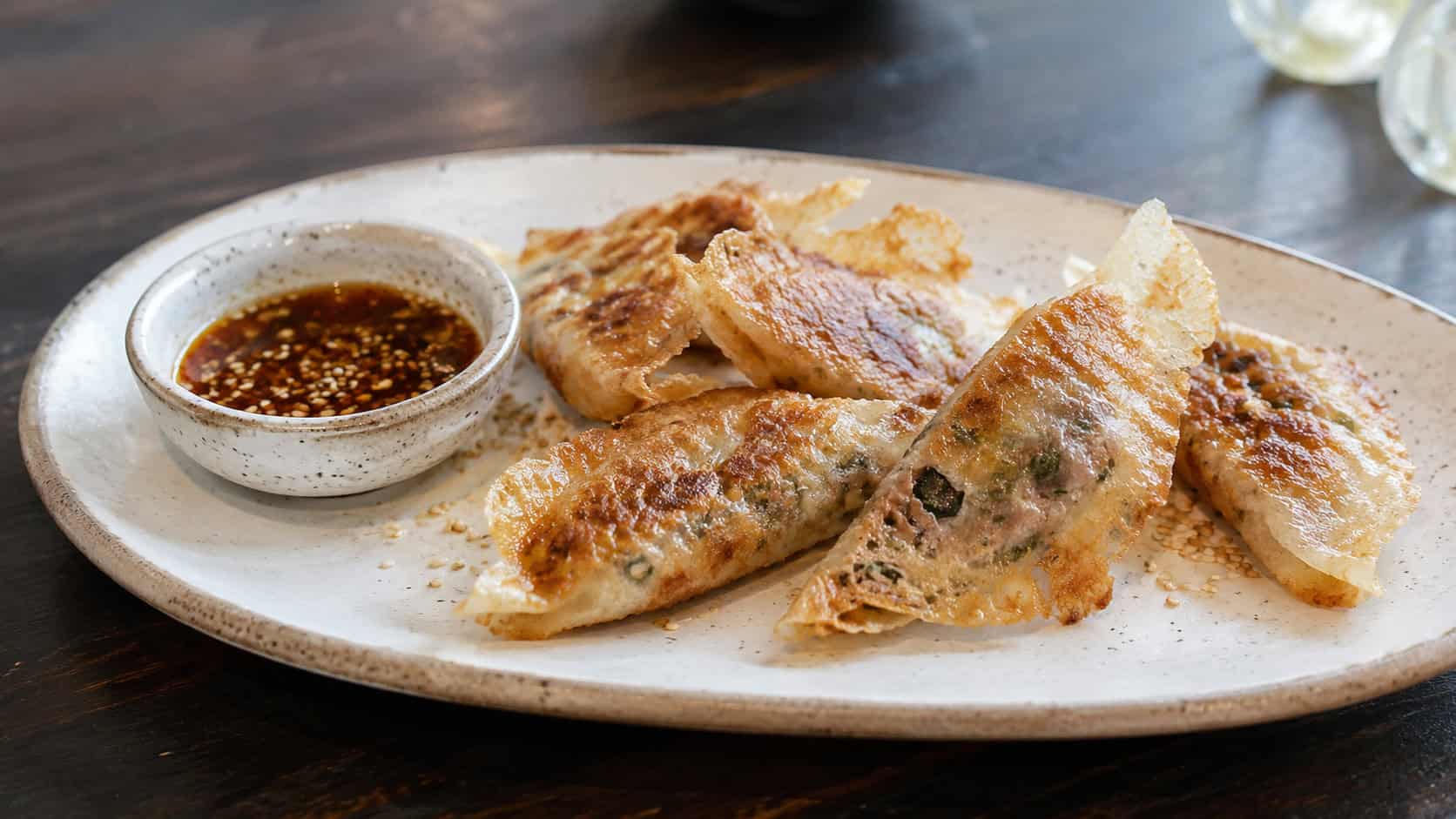 crispy golden rice paper dumplings on a wide plate with a small dish of dipping sauce and sesame seeds, in a wide horizontal shot, on a dark wood table, natural light, shallow depth of field, landscape orientation