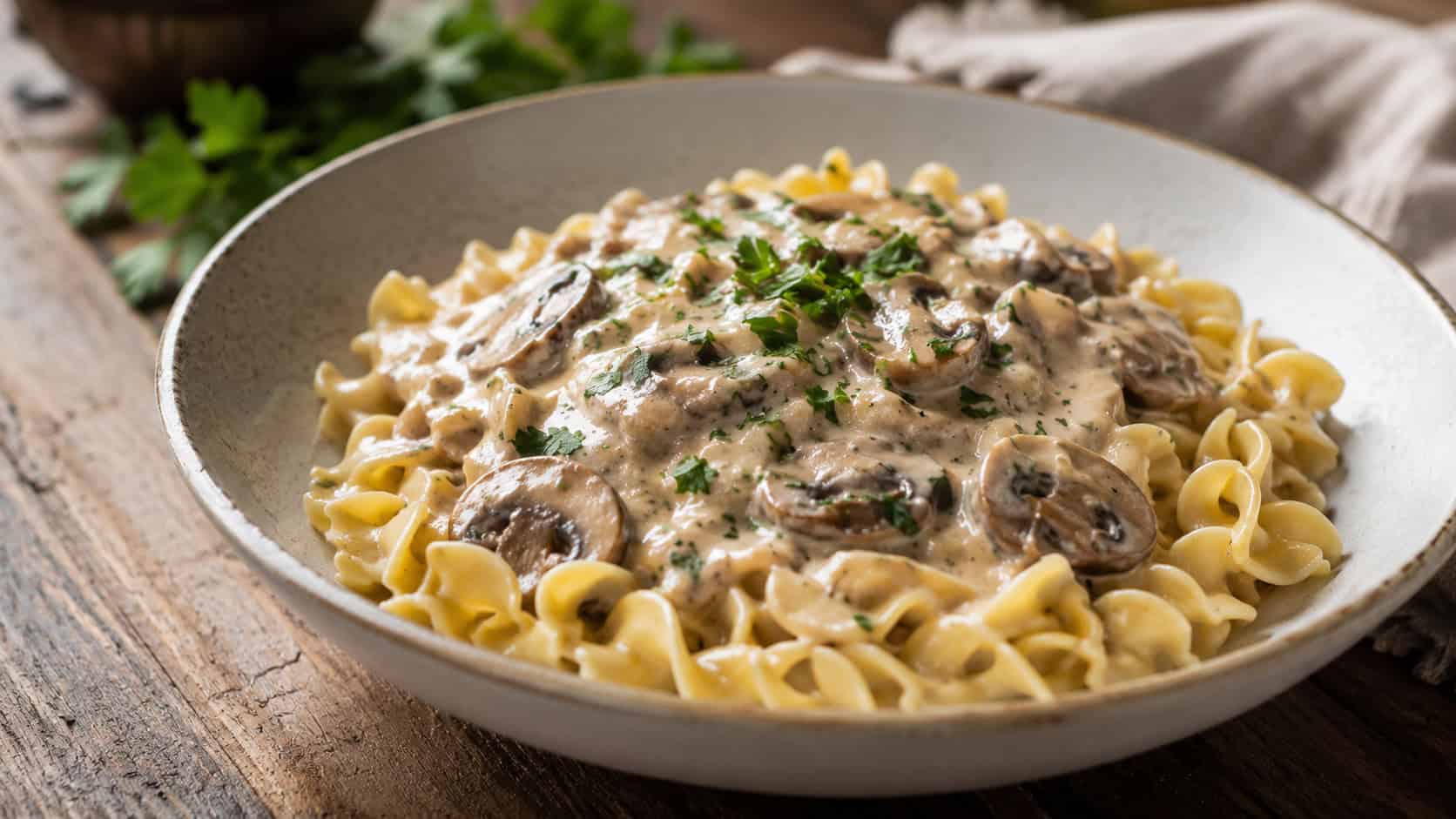 creamy mushroom stroganoff over egg noodles in a wide bowl garnished with fresh parsley, in a wide horizontal shot, on a rustic table, natural light, shallow depth of field, landscape orientation