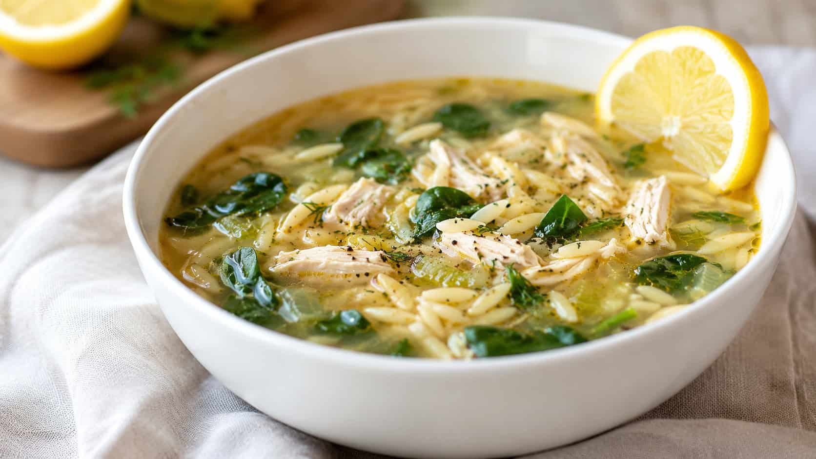 lemon chicken orzo soup in a white bowl, in a wide horizontal shot, with spinach and a lemon slice on the rim, on a soft linen napkin, bright natural light, shallow depth of field, landscape orientation
