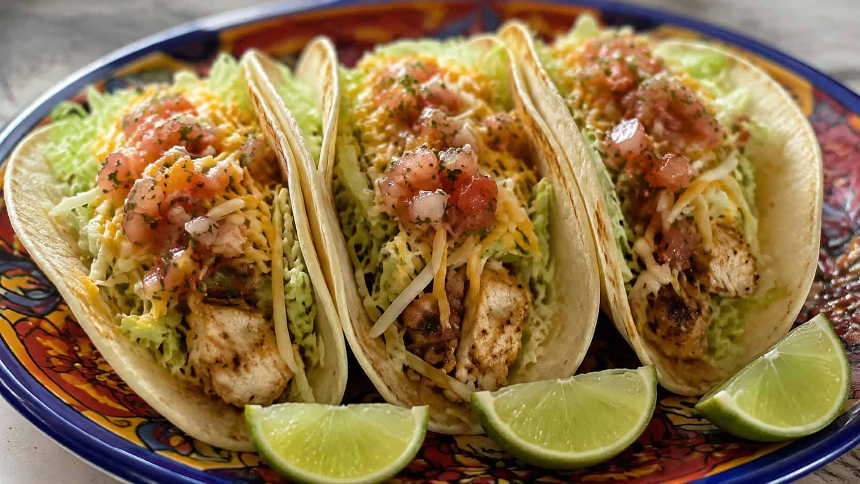 three chicken tacos lined up in a row, in a wide horizontal shot, topped with lettuce, cheese, and salsa, on a colorful plate with lime wedges, natural light, shallow depth of field, landscape orientation