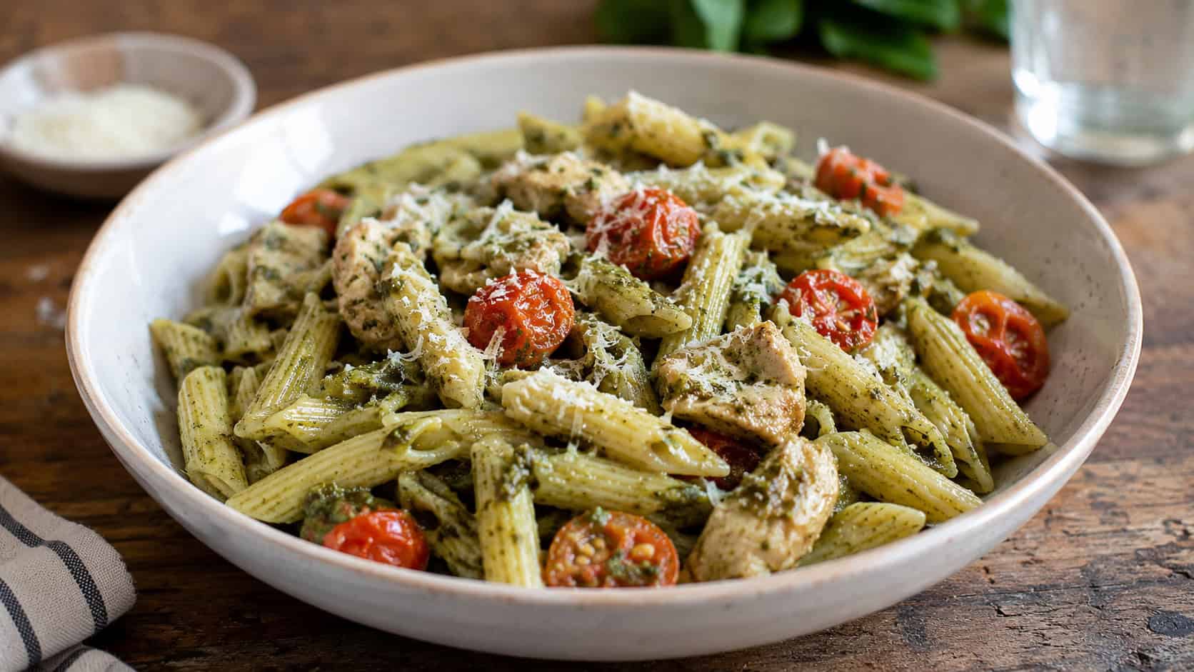 chicken pesto penne with cherry tomatoes in a wide bowl, in a wide horizontal shot, green pesto coating the pasta, parmesan on top, on a rustic table, natural light, shallow depth of field, landscape orientation