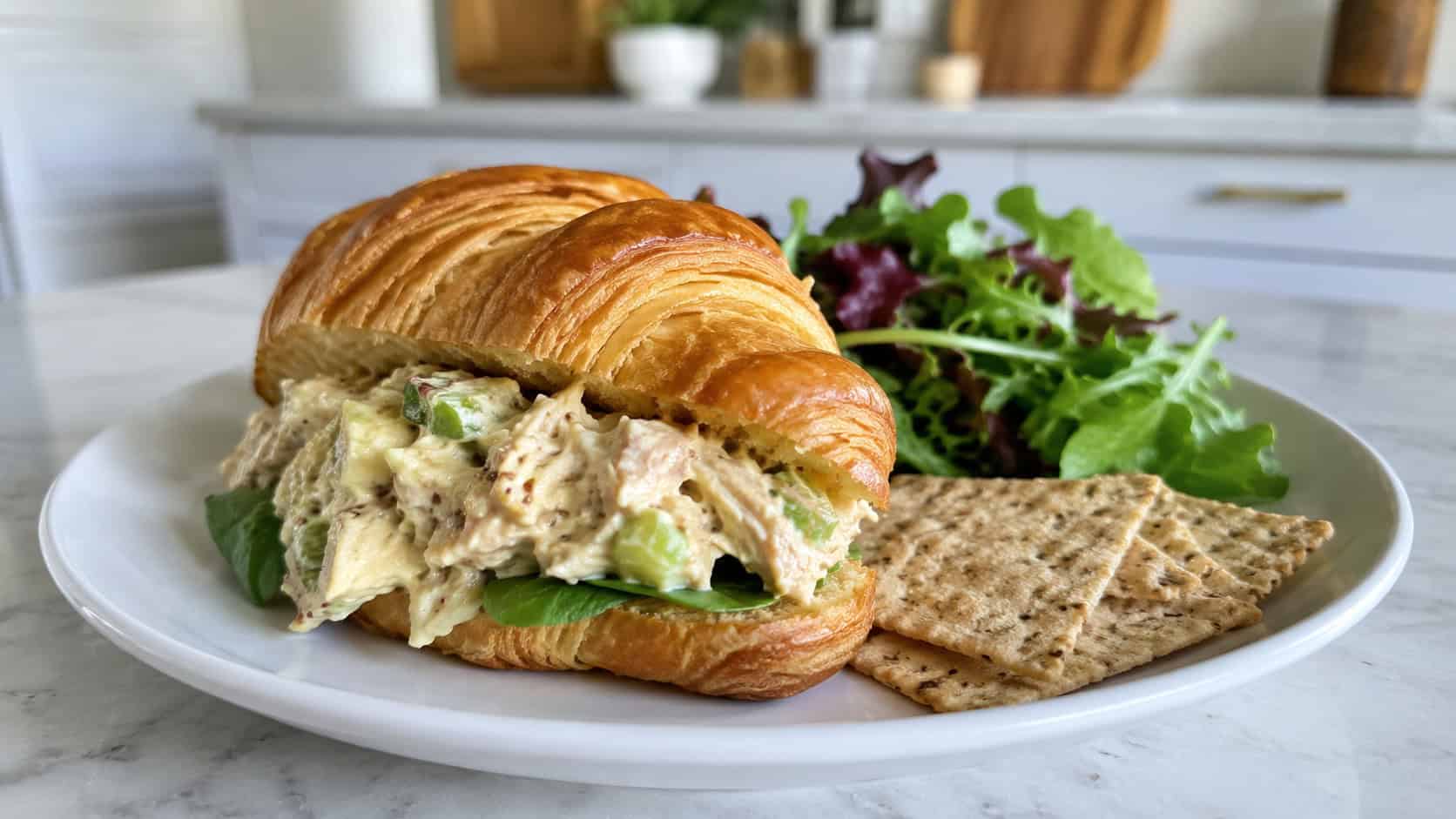 honey mustard chicken salad scooped onto a croissant, in a wide horizontal shot, with greens and crackers on the plate, on a bright kitchen counter, natural light, shallow depth of field, landscape orientation