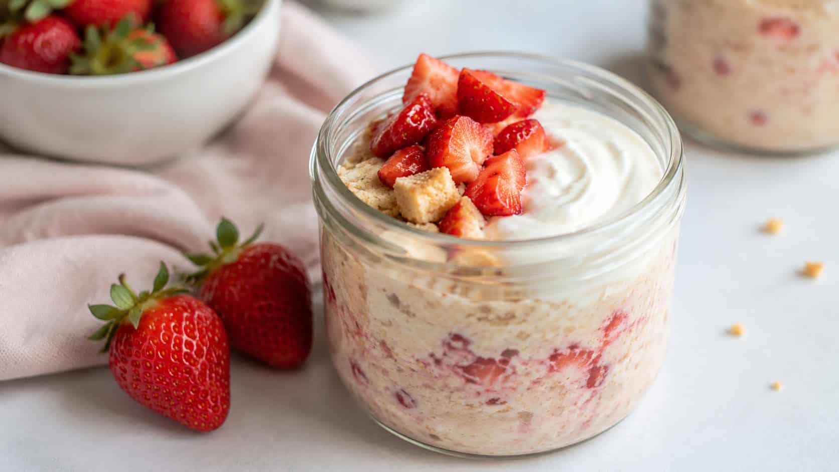 a wide glass jar of strawberry shortcake overnight oats topped with diced strawberries and a swirl of creamy yogurt, fresh strawberries beside it, on a bright table, natural light, shallow depth of field, landscape orientation