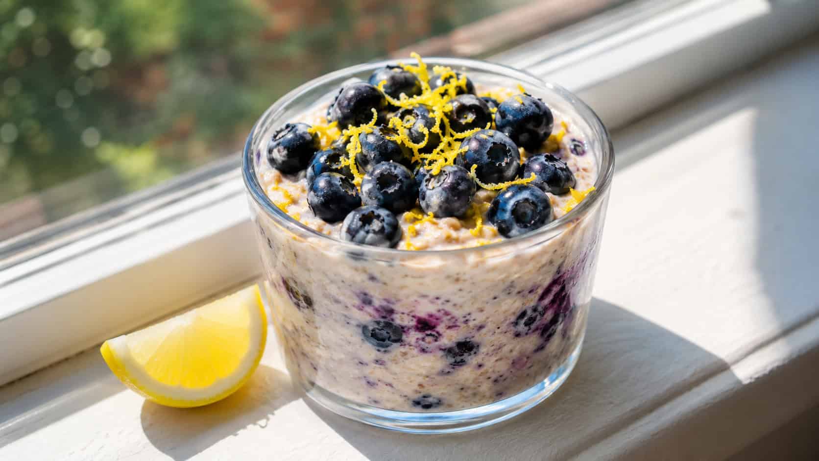 a wide glass jar of lemon blueberry overnight oats topped with plump blueberries and bright lemon zest, a lemon wedge beside it, on a sunny windowsill, bright natural light, shallow depth of field, landscape orientation