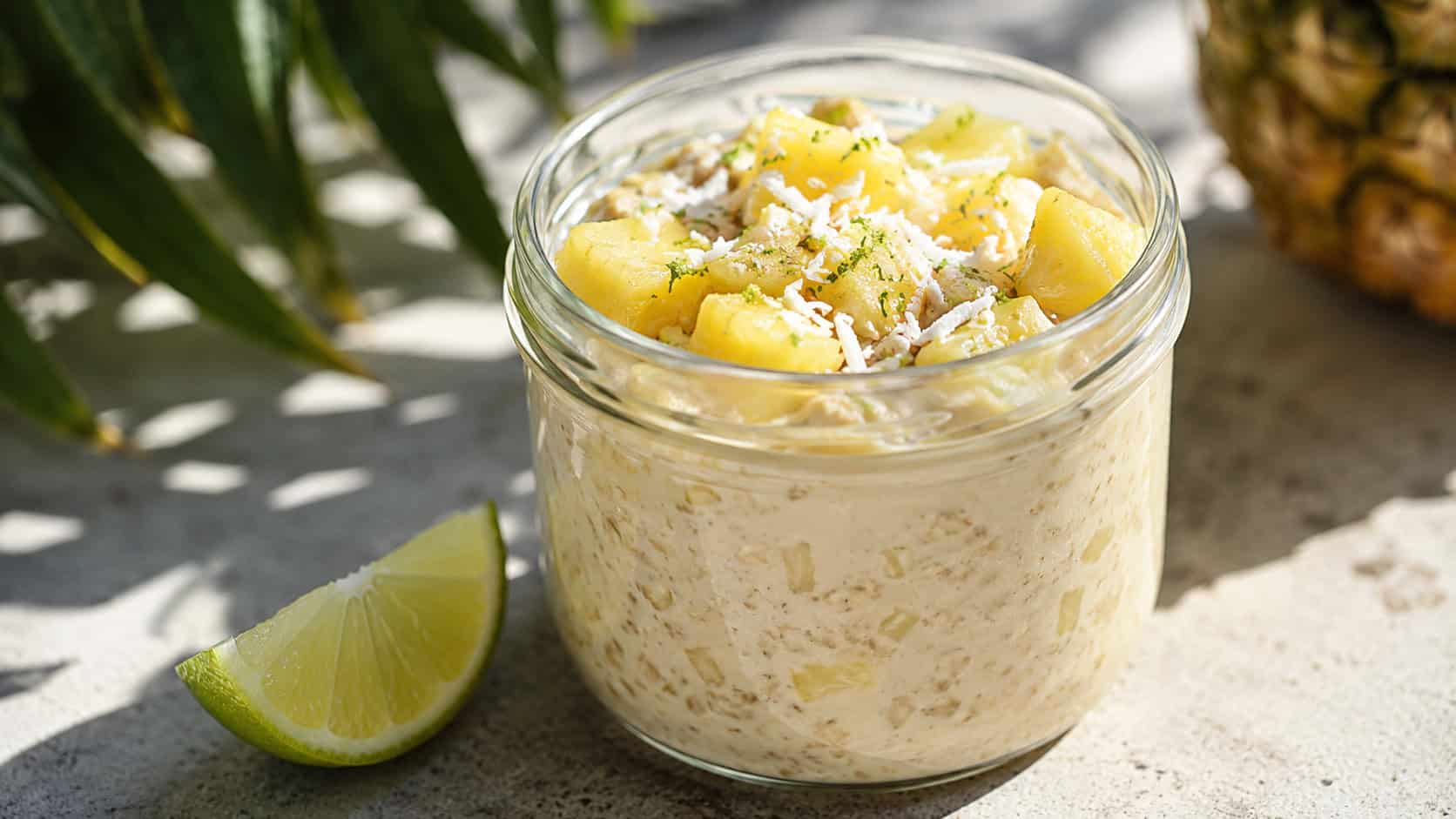 a wide glass jar of tropical pineapple overnight oats topped with golden pineapple chunks, shredded coconut, and lime zest, a lime wedge beside it, on a bright sunny surface, natural light, shallow depth of field, landscape orientation