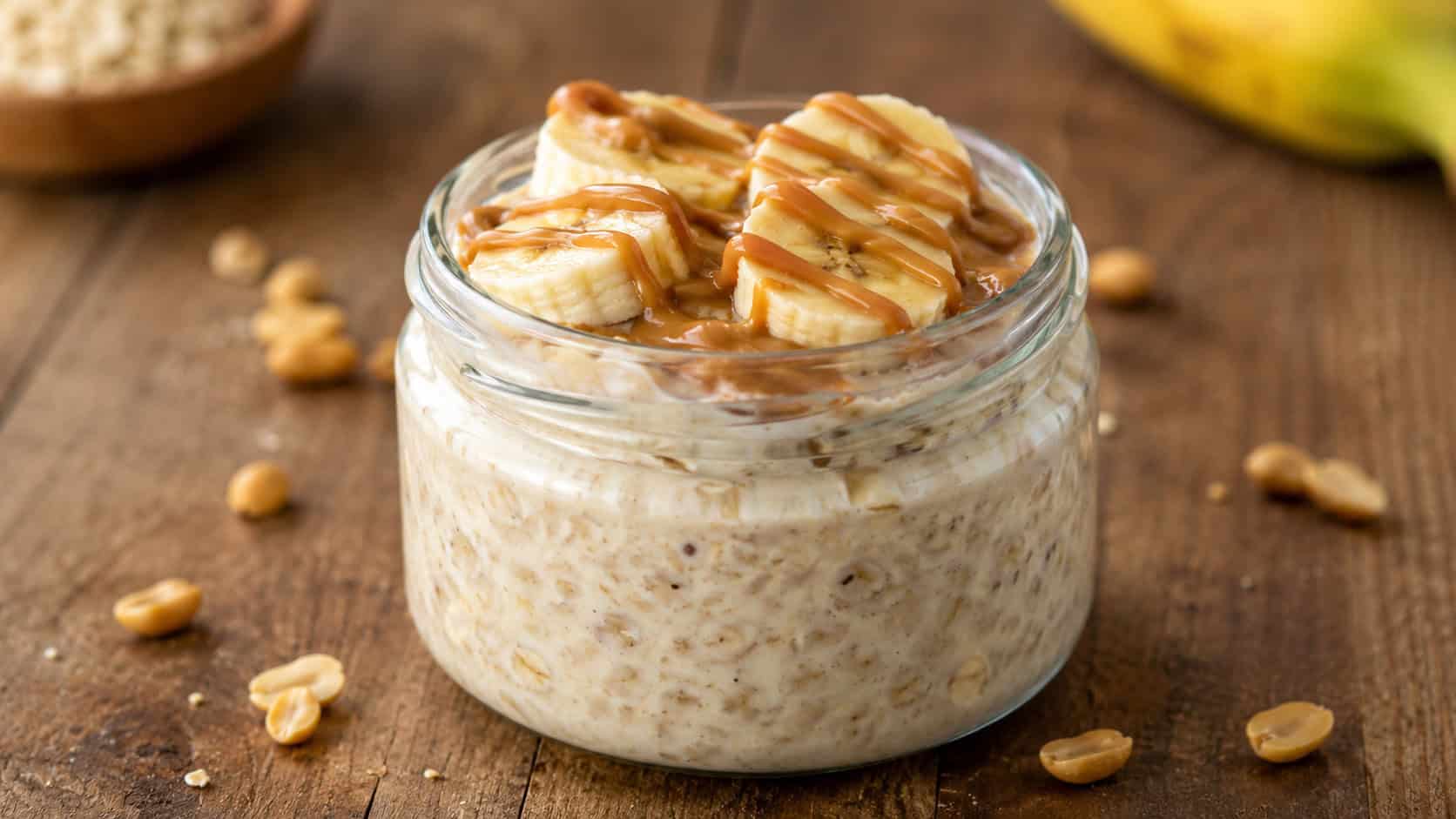 a wide glass jar of peanut butter banana overnight oats topped with banana slices and a peanut butter drizzle, a few peanuts scattered around, on a wooden table, warm natural light, shallow depth of field, landscape orientation