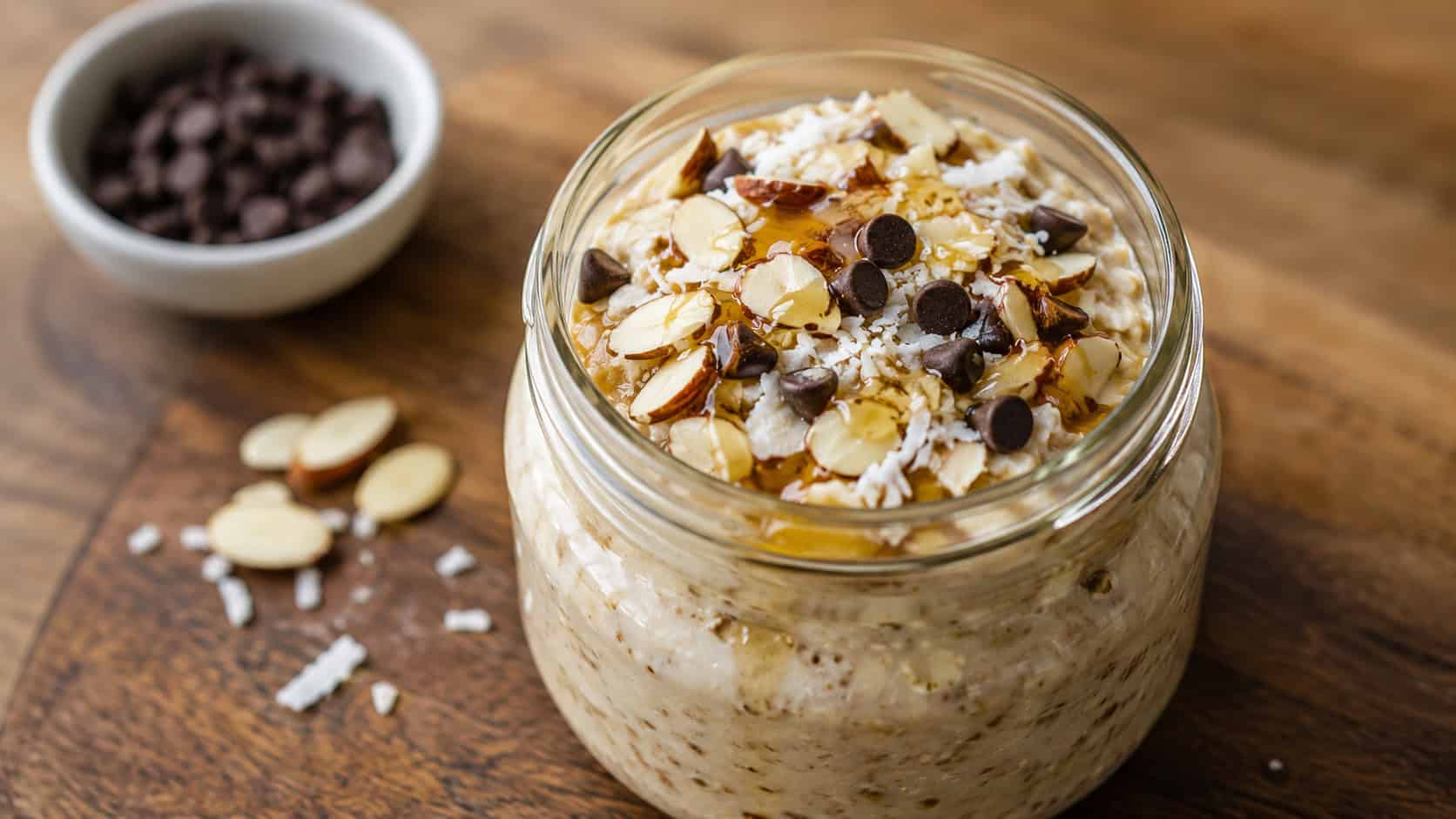 a wide glass jar of honey almond overnight oats topped with sliced almonds, shredded coconut, and chocolate chips, a honey drizzle on top, on a wooden board, warm natural light, shallow depth of field, landscape orientation