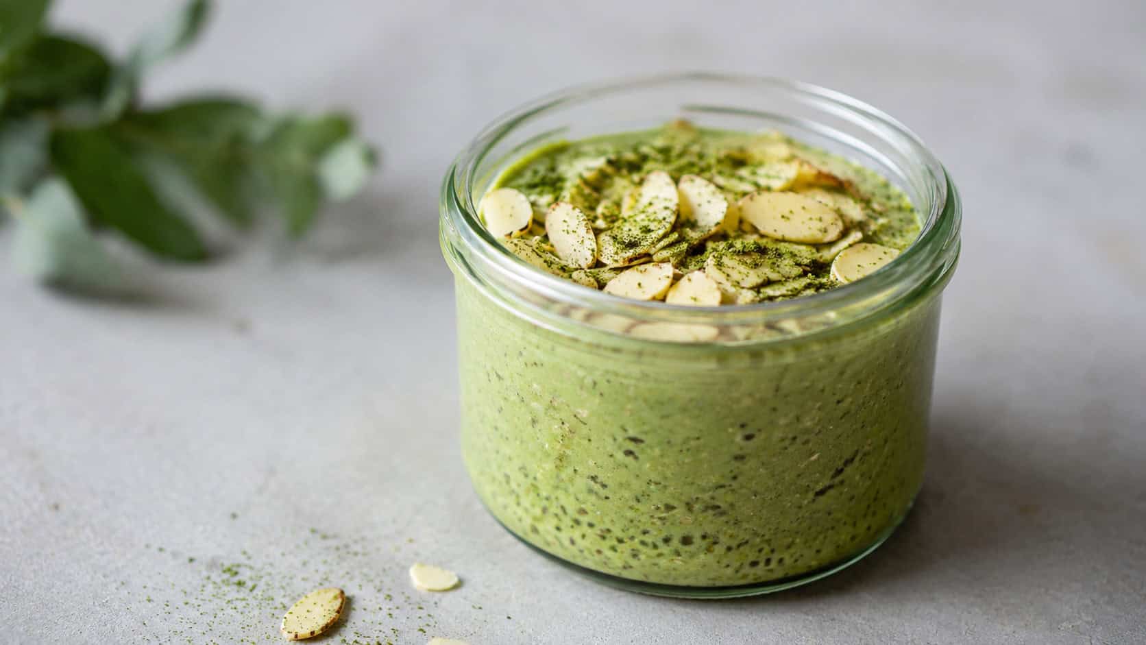 a wide glass jar of vibrant green matcha overnight oats topped with sliced almonds and a light dusting of matcha powder, on a minimalist surface, soft natural light, shallow depth of field, landscape orientation
