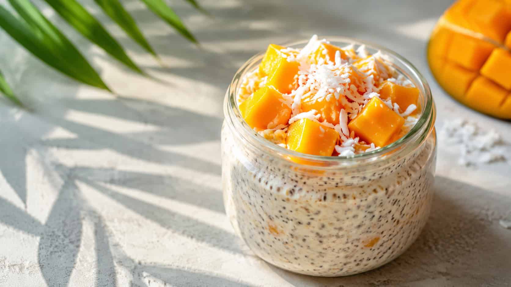 a wide glass jar of coconut overnight oats topped with bright orange mango chunks and shredded coconut, on a light tropical surface with a palm shadow, bright natural light, shallow depth of field, landscape orientation