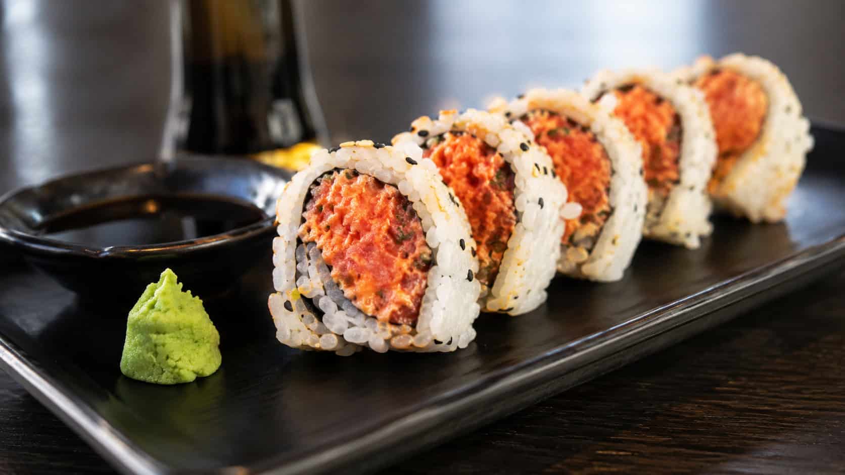 spicy tuna sushi rolls in a wide horizontal shot, neat rounds showing pink tuna and white rice with a drizzle of spicy mayo, arranged on a black slate with wasabi and ginger, bright natural light, shallow depth of field, landscape orientation