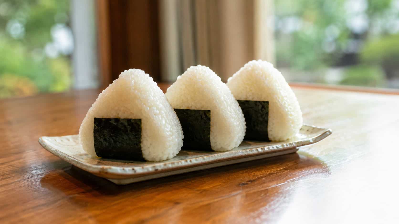 rice triangle onigiri in a wide horizontal shot, neat white rice wrapped with dark nori strips and sprinkled with sesame seeds, arranged on a wooden board with a small bowl of soy sauce, bright natural light, shallow depth of field, landscape orientation