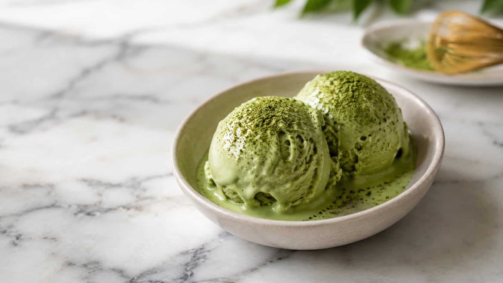 scoops of pale green matcha ice cream in a wide horizontal shot, melting slightly in a shallow bowl dusted with matcha powder, a small spoon alongside on a light marble surface, soft bright light, shallow depth of field, landscape orientation