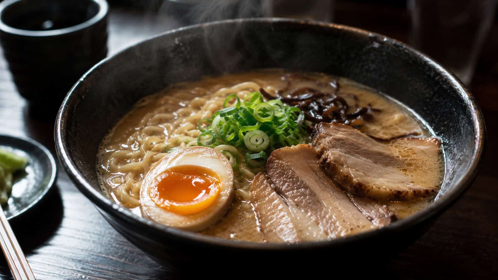 a bowl of pork ramen in a wide horizontal shot, curly noodles in deep broth topped with sliced pork, a halved soft egg, green onion, and nori, chopsticks on the rim, set on a dark table, steam rising, moody warm light, shallow depth of field, landscape orientation