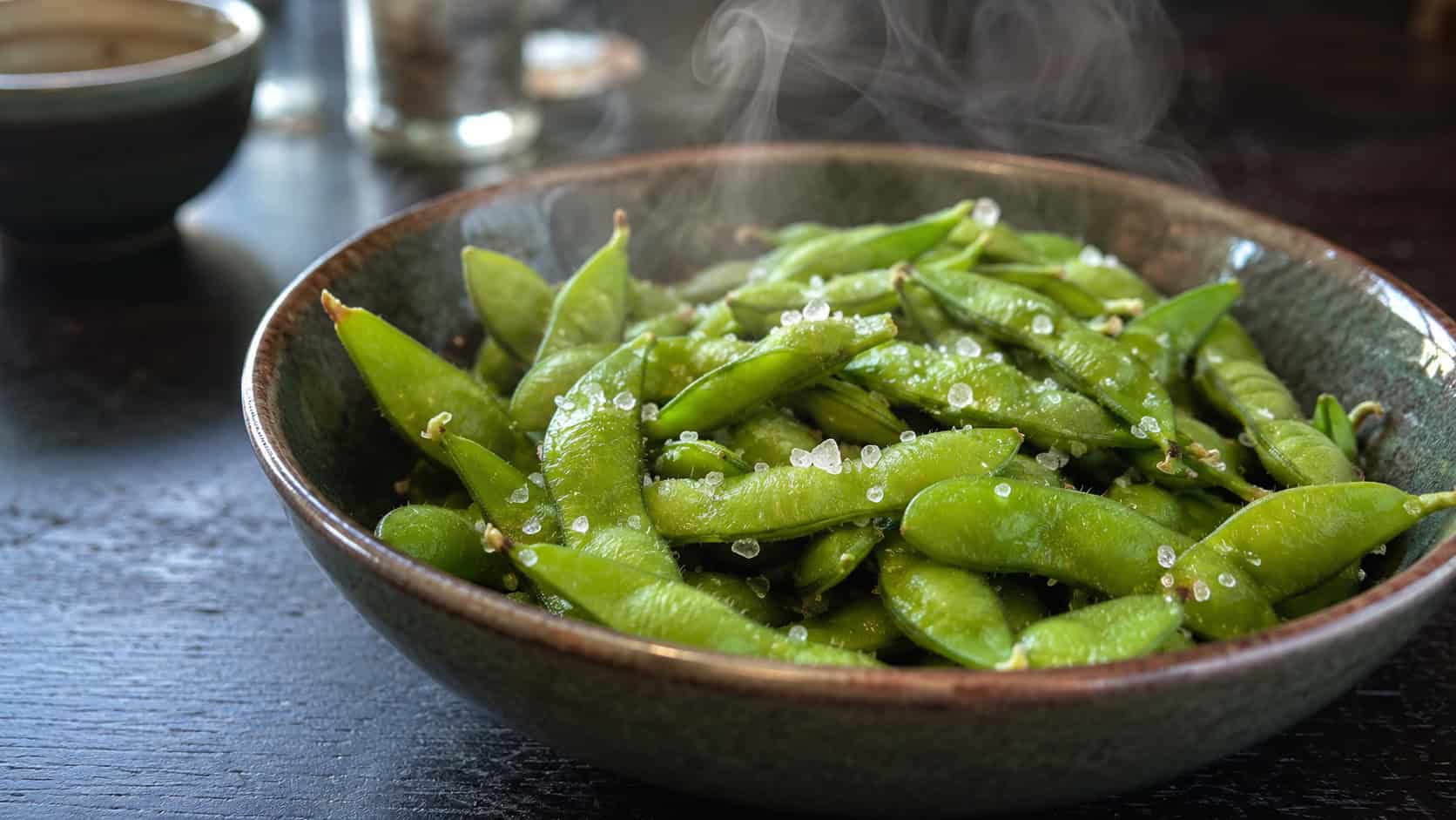 a bowl of bright green edamame pods in a wide horizontal shot, glistening and sprinkled with flaky sea salt, an empty dish for shells alongside on a dark wood table, fresh natural light, shallow depth of field, landscape orientation