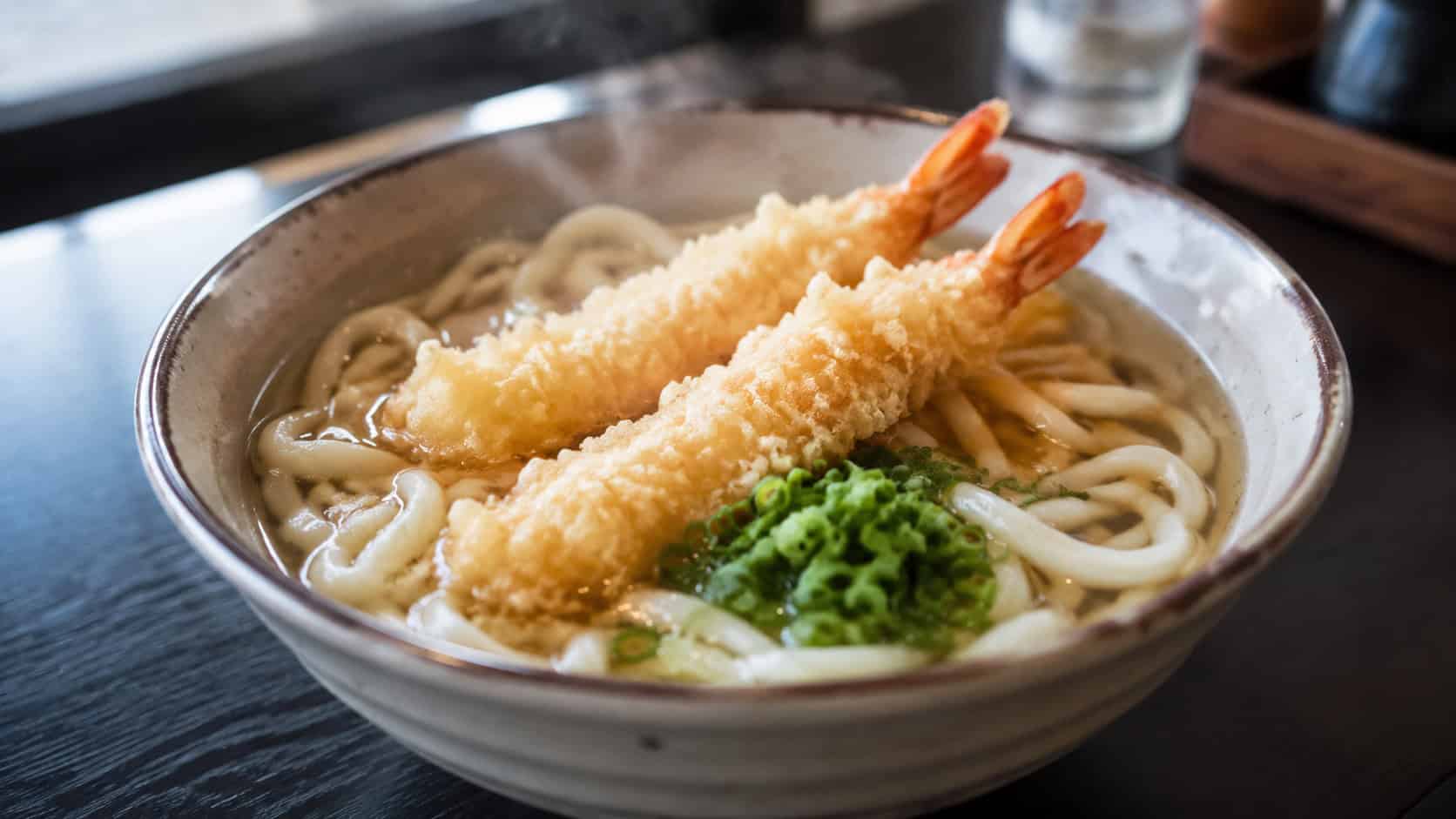 a bowl of tempura udon in a wide horizontal shot, thick noodles in golden broth topped with crispy shrimp tempura and green onion, chopsticks resting across the rim on a dark table, steam rising, warm light, shallow depth of field, landscape orientation