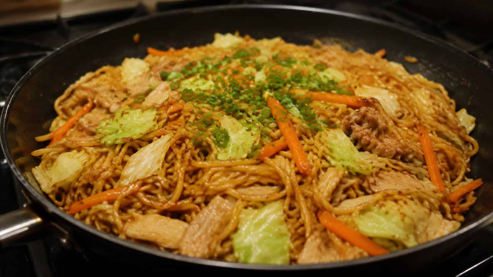 stir-fried yakisoba noodles in a wide horizontal shot, glossy noodles tangled with cabbage, carrot, and pork, garnished with green onion in a black bowl, set on a wooden table, warm light, shallow depth of field, landscape orientation