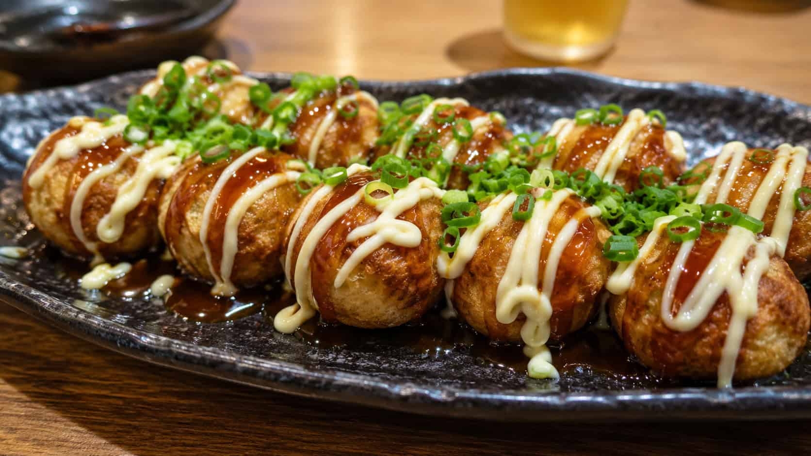 a plate of takoyaki balls in a wide horizontal shot, drizzled with dark sauce and topped with dancing bonito flakes and green onion, arranged on a wooden tray, steam rising, warm street-food light, shallow depth of field, landscape orientation