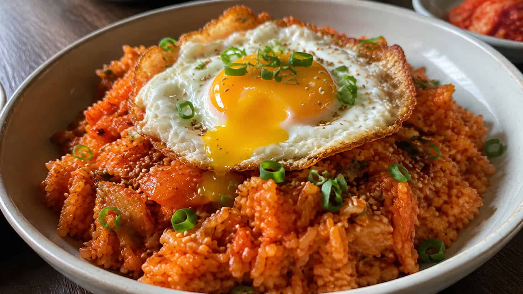 spicy red kimchi fried rice topped with a crispy fried egg with runny yolk in a wide horizontal shot in a bowl, sprinkled with sesame seeds and green onions, natural light, shallow depth of field, landscape orientation