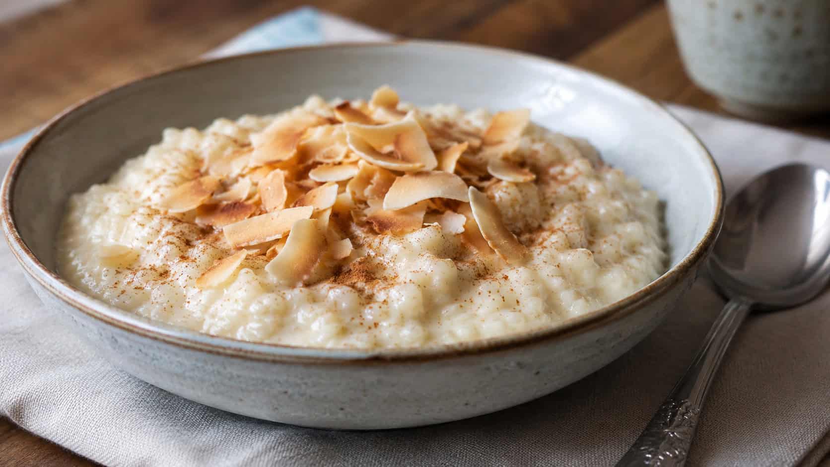 creamy coconut rice pudding in a wide horizontal shot in a shallow bowl, dusted with cinnamon and topped with toasted coconut flakes, a spoon resting beside it, soft natural light, shallow depth of field, landscape orientation