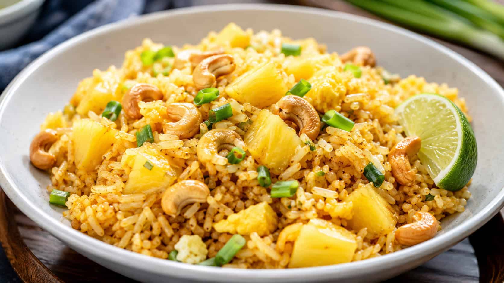 golden Thai pineapple fried rice with juicy pineapple chunks, cashews, and green onions in a wide horizontal shot in a bowl, a lime wedge on the side, bright natural light, shallow depth of field, landscape orientation