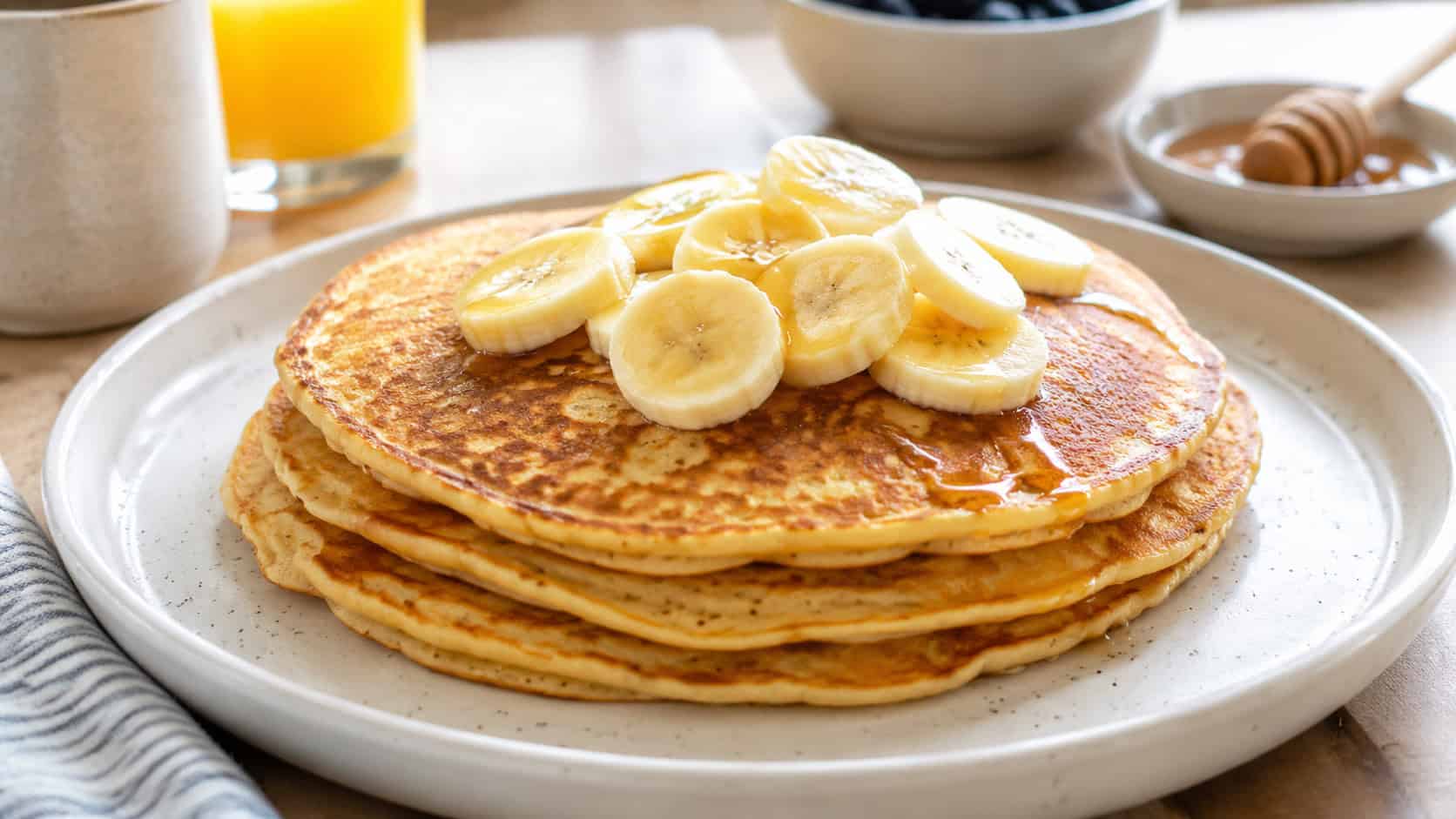 a wide plate of golden banana pancakes stacked with sliced banana on top, on a bright breakfast table, natural light, shallow depth of field, landscape orientation