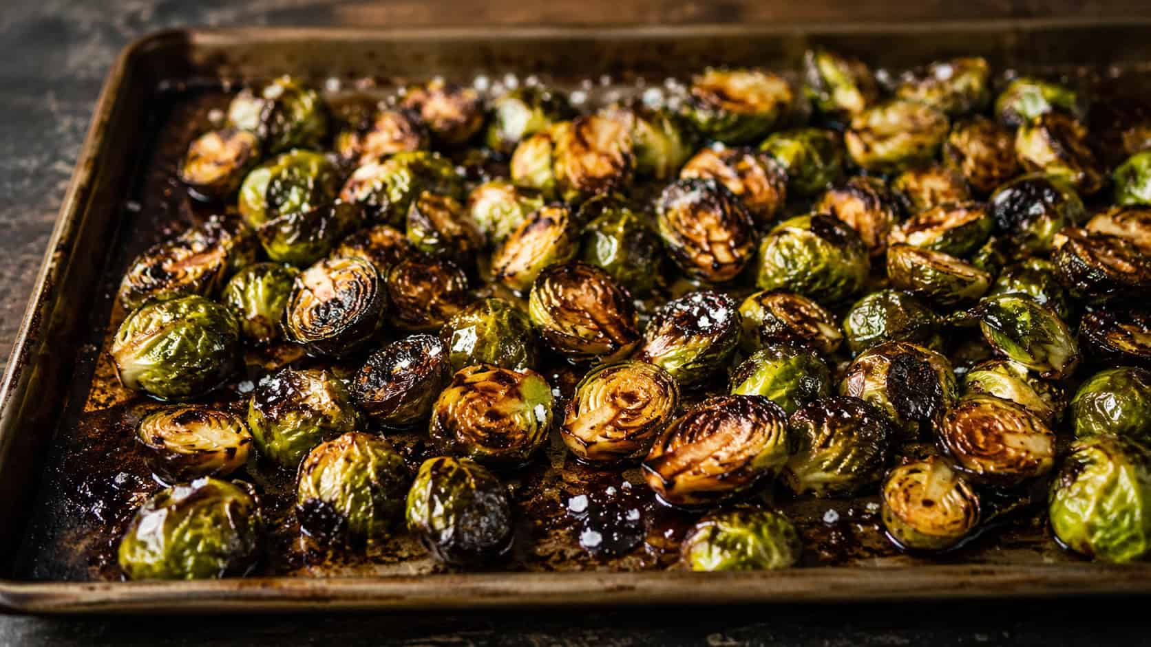 balsamic glazed Brussels sprouts on a wide sheet pan shot horizontally, crispy charred sprouts coated in glossy dark glaze, a sprinkle of flaky salt on a rustic dark surface, warm natural light, shallow depth of field, landscape orientation
