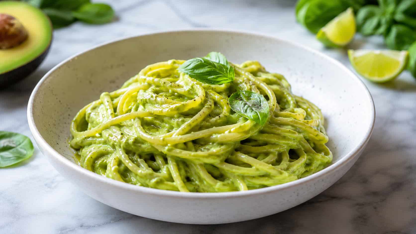 creamy green avocado pasta twirled in a wide bowl shot horizontally, glossy linguine in bright basil-avocado sauce, lime wedges and fresh basil leaves on a light marble surface, natural light, shallow depth of field, landscape orientation