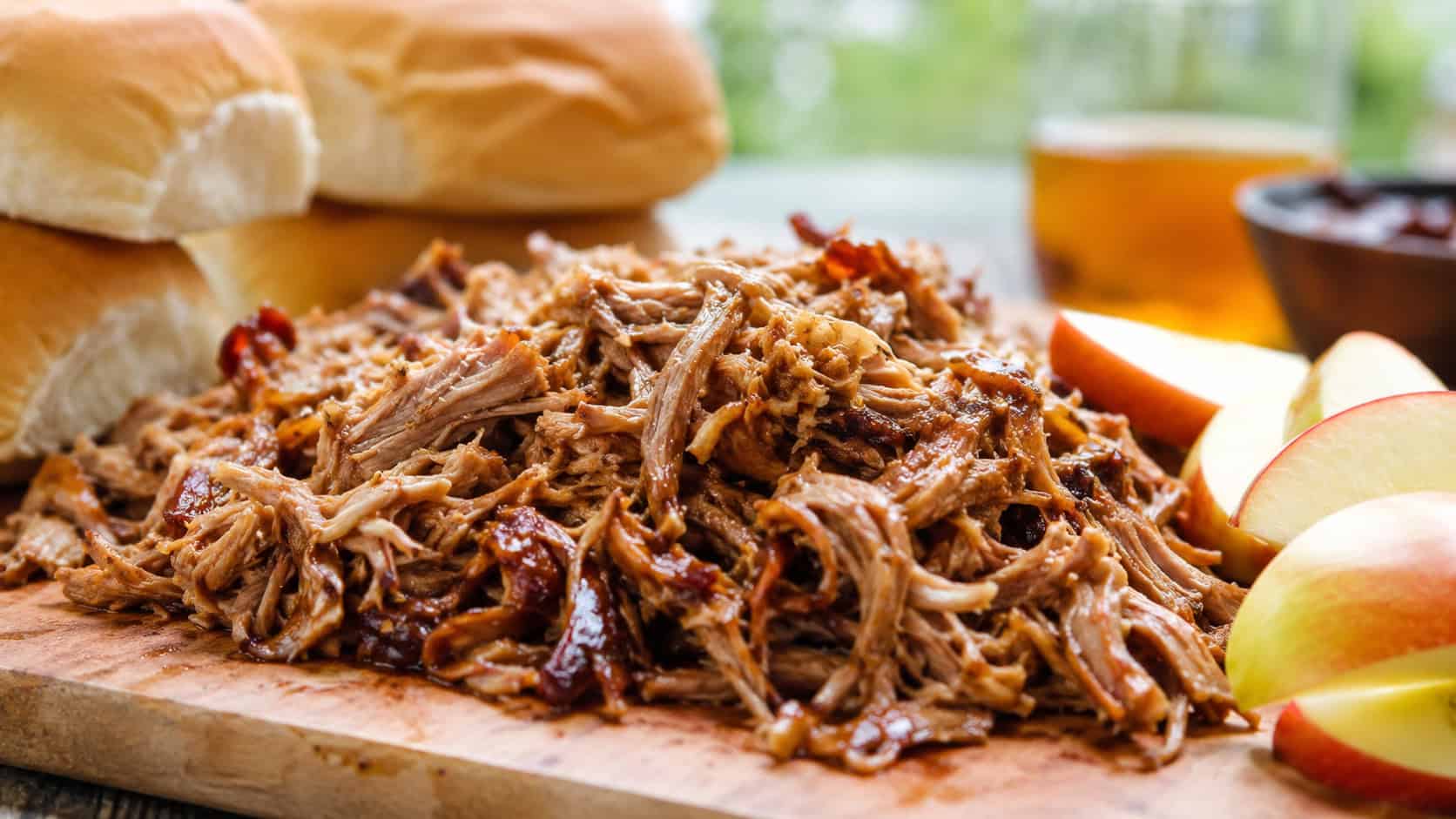 tender shredded apple cider pulled pork in a wide horizontal shot, piled on a board with soft buns and sliced apples nearby, natural light, shallow depth of field, landscape orientation