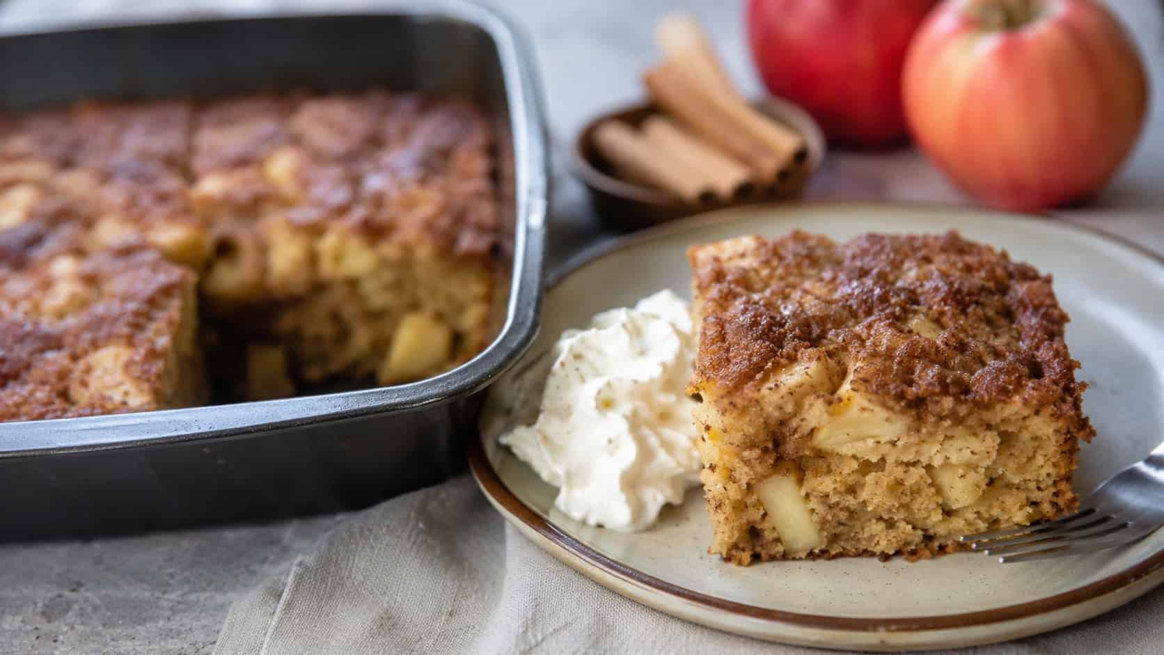 a warm cinnamon-sugar apple cake in a wide horizontal shot, one square slice lifted from the pan with whipped cream beside it, natural light, shallow depth of field, landscape orientation