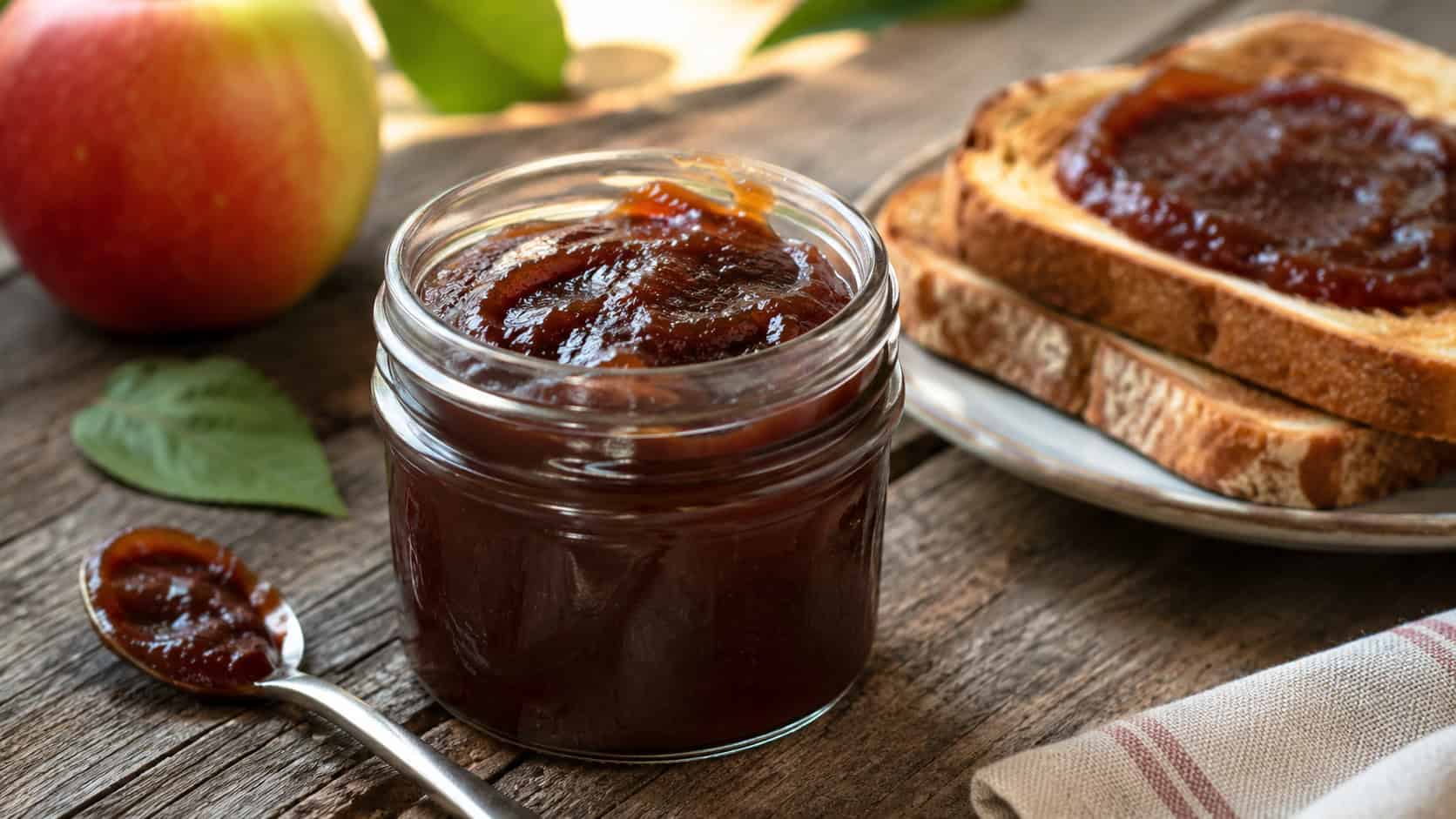 a jar of thick glossy dark apple butter in a wide horizontal shot, with a spoon and slices of toast on a rustic wooden table, natural light, shallow depth of field, landscape orientation