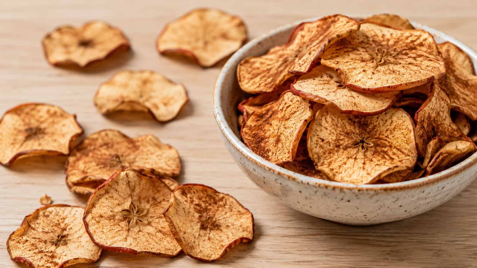 thin crispy cinnamon apple chips in a wide horizontal shot, piled in a bowl and scattered across a light wooden surface, natural light, shallow depth of field, landscape orientation