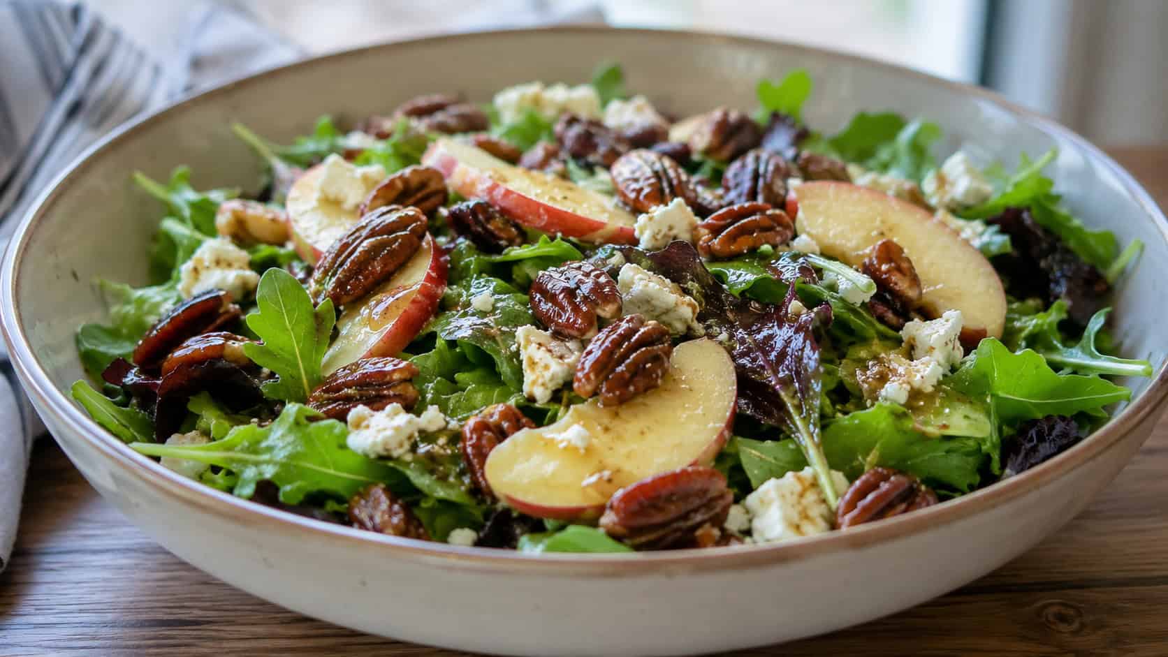 a fresh green salad with apple slices, candied pecans, and goat cheese in a wide horizontal shot, in a wide serving bowl drizzled with vinaigrette, natural light, shallow depth of field, landscape orientation