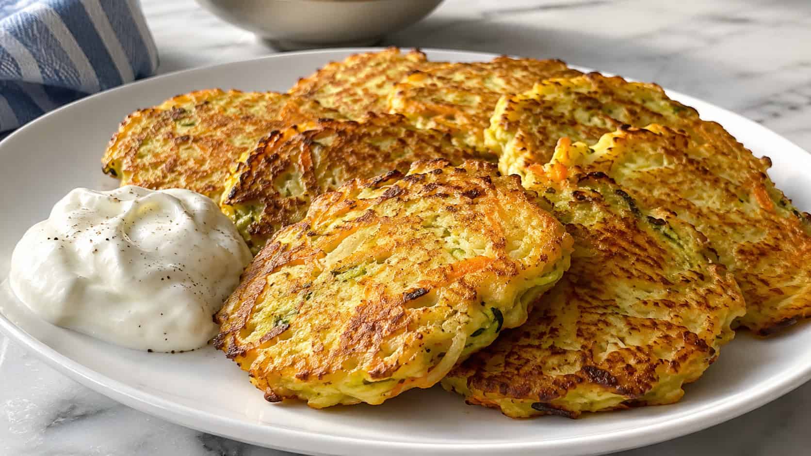 a wide plate of golden savory vegetable pancakes flecked with carrot and zucchini, a dollop of sour cream on the side, on a bright table, natural light, shallow depth of field, landscape orientation