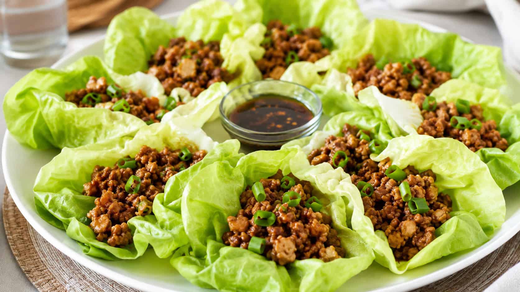 a wide platter of Asian chicken lettuce wraps with crisp green butter lettuce cups filled with savory seasoned ground chicken and green onion, on a bright table, natural light, shallow depth of field, landscape orientation