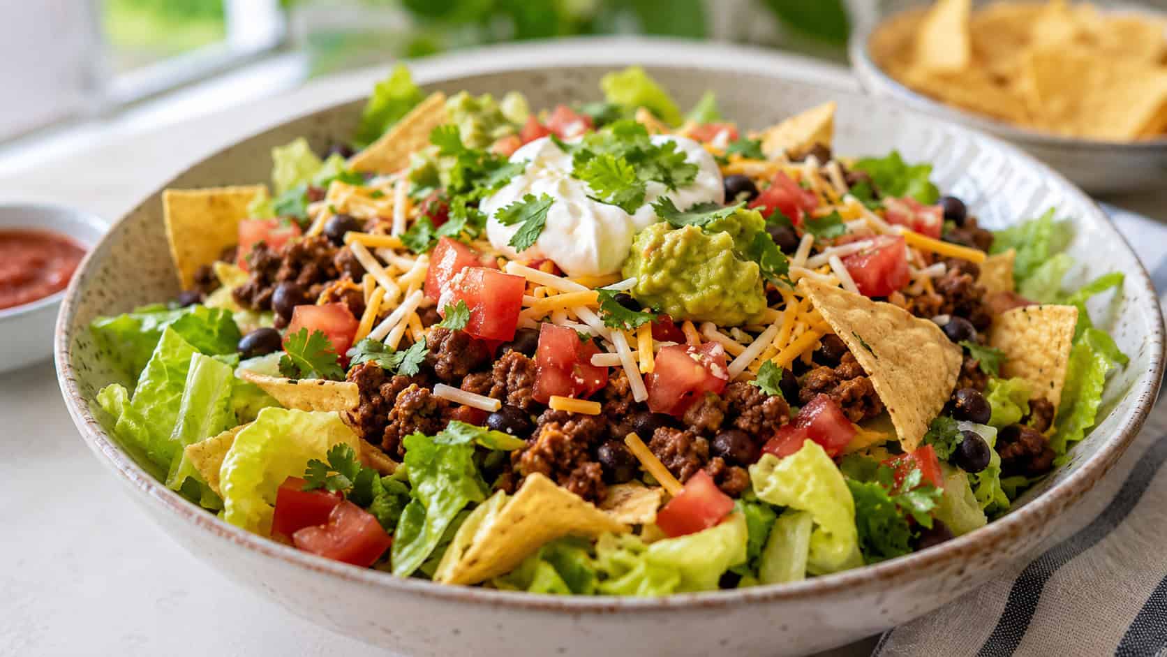 a wide colorful taco salad bowl with crisp lettuce, seasoned ground beef, shredded cheese, tomatoes, black beans and tortilla chips, on a bright table, natural light, shallow depth of field, landscape orientation