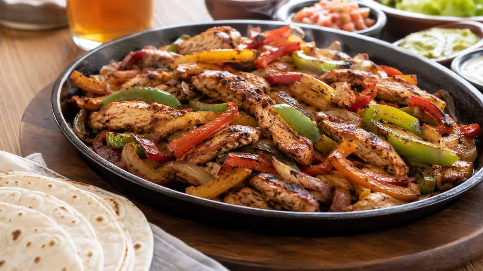 a wide sizzling skillet of chicken fajitas with charred bell peppers, onions and seasoned chicken strips, warm tortillas alongside, on a wooden table, natural light, shallow depth of field, landscape orientation