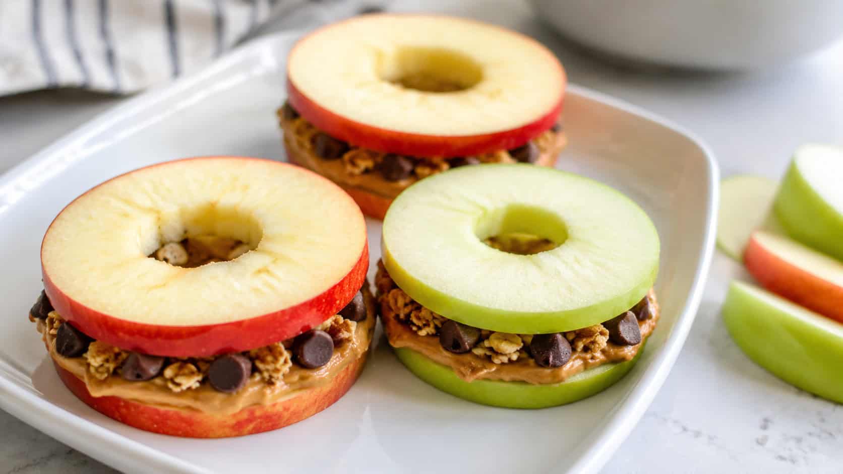 apple ring sandwiches filled with peanut butter and granola on a plate in a wide horizontal composition, chocolate chips peeking out, fresh apple slices nearby on a bright table, natural light, shallow depth of field, landscape orientation