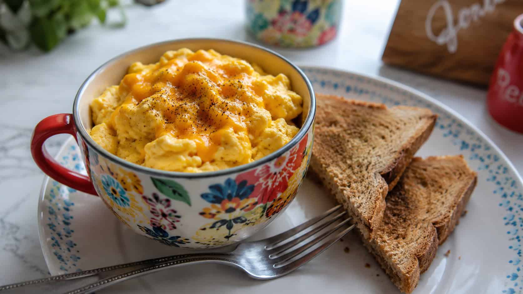 fluffy scrambled eggs in a colorful mug in a wide horizontal composition, melty cheese on top, a fork and toast triangles beside it on a bright kitchen table, natural light, shallow depth of field, landscape orientation
