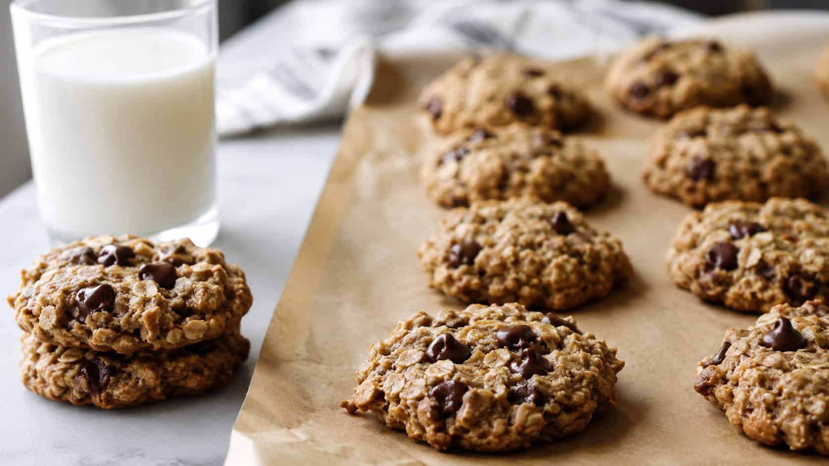 soft banana oat cookies cooling on parchment in a wide horizontal shot, melty chocolate chips and rustic oat texture, a couple stacked beside a glass of milk on a bright table, natural light, shallow depth of field, landscape orientation
