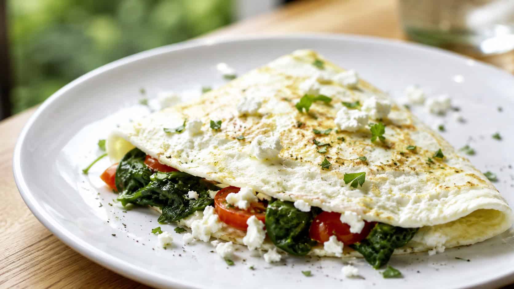 a folded egg white omelette on a white plate in a wide horizontal shot, spinach and tomato peeking out, crumbled feta, fresh herbs, bright natural light, shallow depth of field, landscape orientation