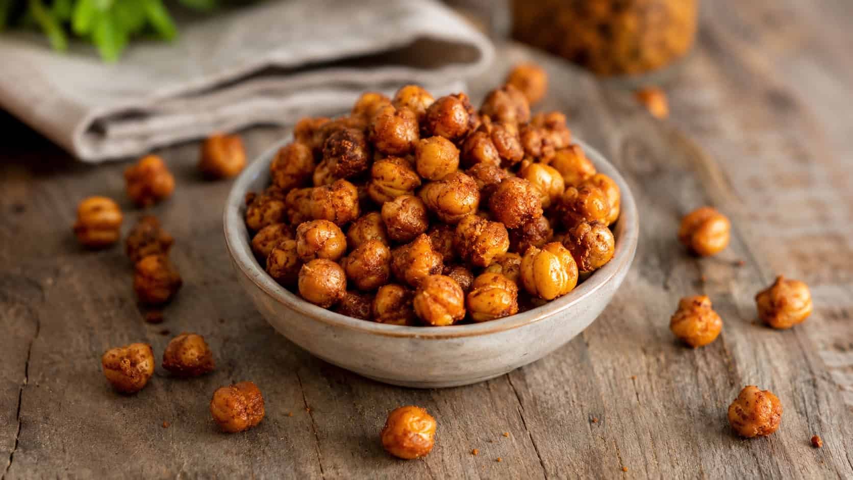 crispy roasted chickpeas in a small bowl in a wide horizontal shot, golden spiced exterior, scattered on a rustic table, warm natural light, shallow depth of field, landscape orientation