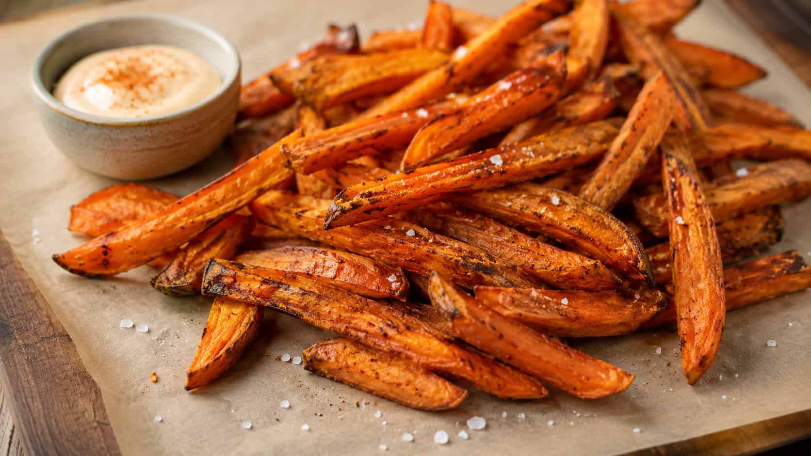 crispy baked sweet potato fries piled on parchment in a wide horizontal shot, orange wedges, small bowl of dip, scattered sea salt, warm natural light, shallow depth of field, landscape orientation