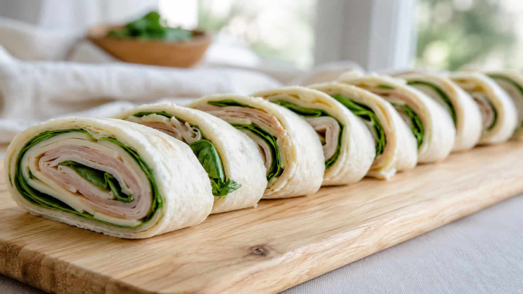a wide horizontal plate of turkey and Swiss tortilla pinwheels arranged in a row, green spinach peeking out, on a light wood board, natural light, shallow depth of field, landscape orientation