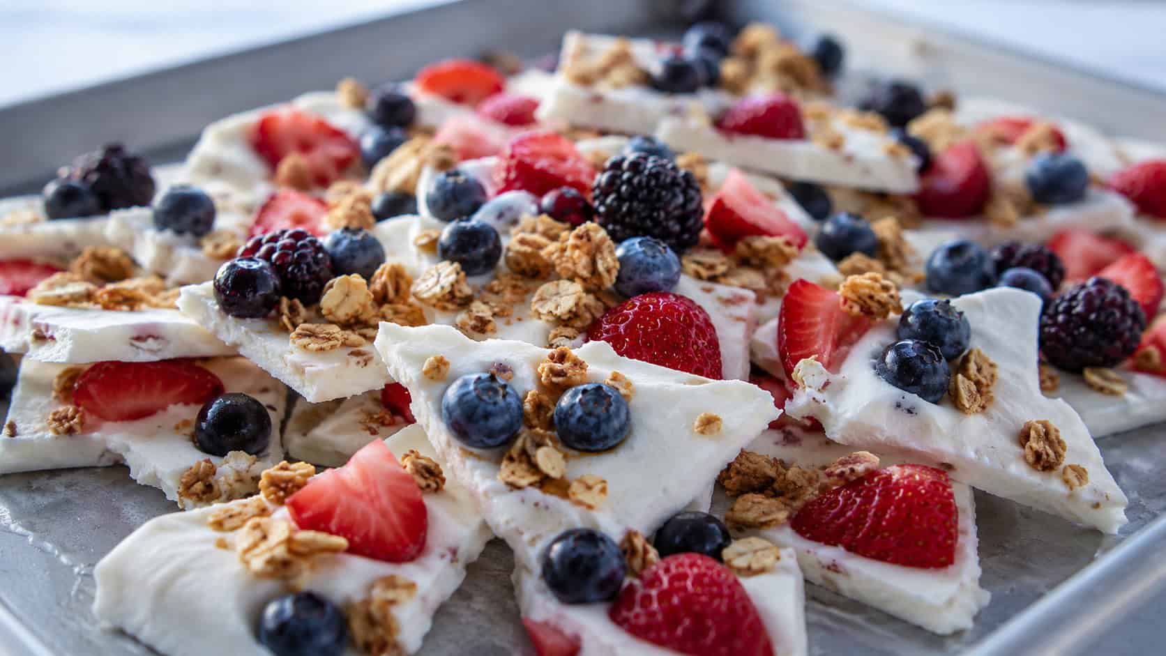 frozen yogurt bark broken into shards topped with berries and granola in a wide horizontal shot, on a chilled metal tray, cool natural light, shallow depth of field, landscape orientation